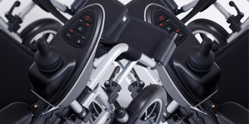 Two power wheelchairs crossing over one another to create an abstract entanglement. The control panels overlap and show red green and white buttons. Both joysticks point forward.