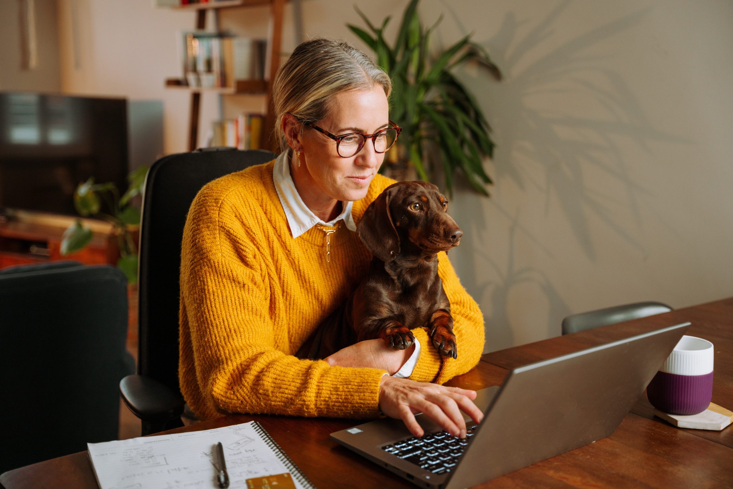 A white woman with blonde hair pulled back into a bun and wearing glasses and a dandelion yellow sweater holds a brown dachshund dog in one arm and types on a laptop with her other hand. The laptop sits on a wooden kitchen table. Beside it is an open notebook. A television and bookshelf is visible behind the woman.