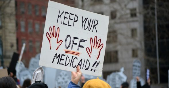 In a crowd, a person wearing a yellow beret holds up a sign that says keep your a drawing of two hands OFF Medicaid!!