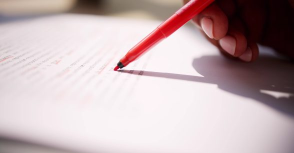 A red pen on a piece on paper underlining blurred out words in red ink. A Black person with short light pink acrylic nails holds the red pen.